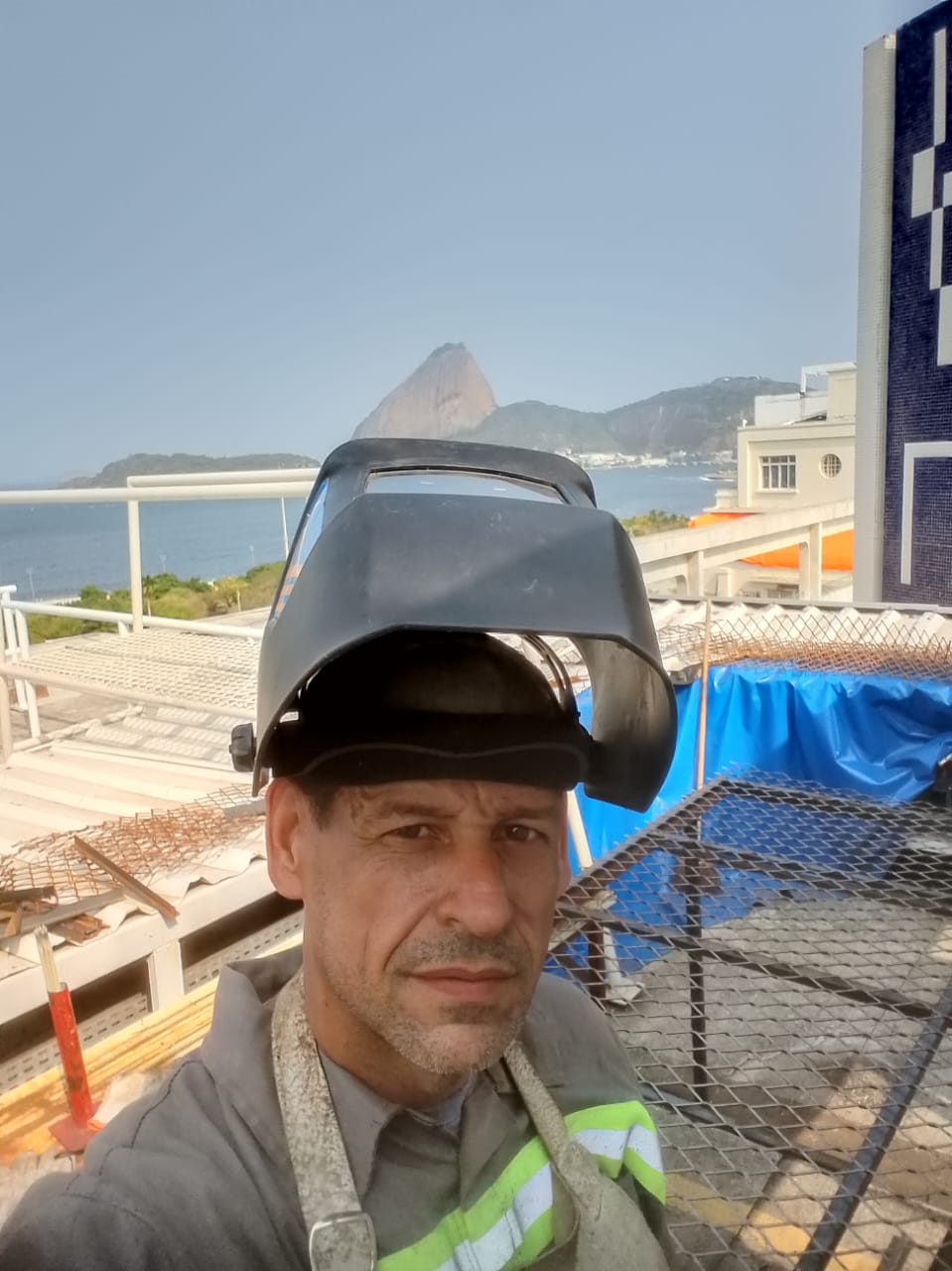 Trabalhador com capacete de soldador em telhado com vista para o morro e o mar